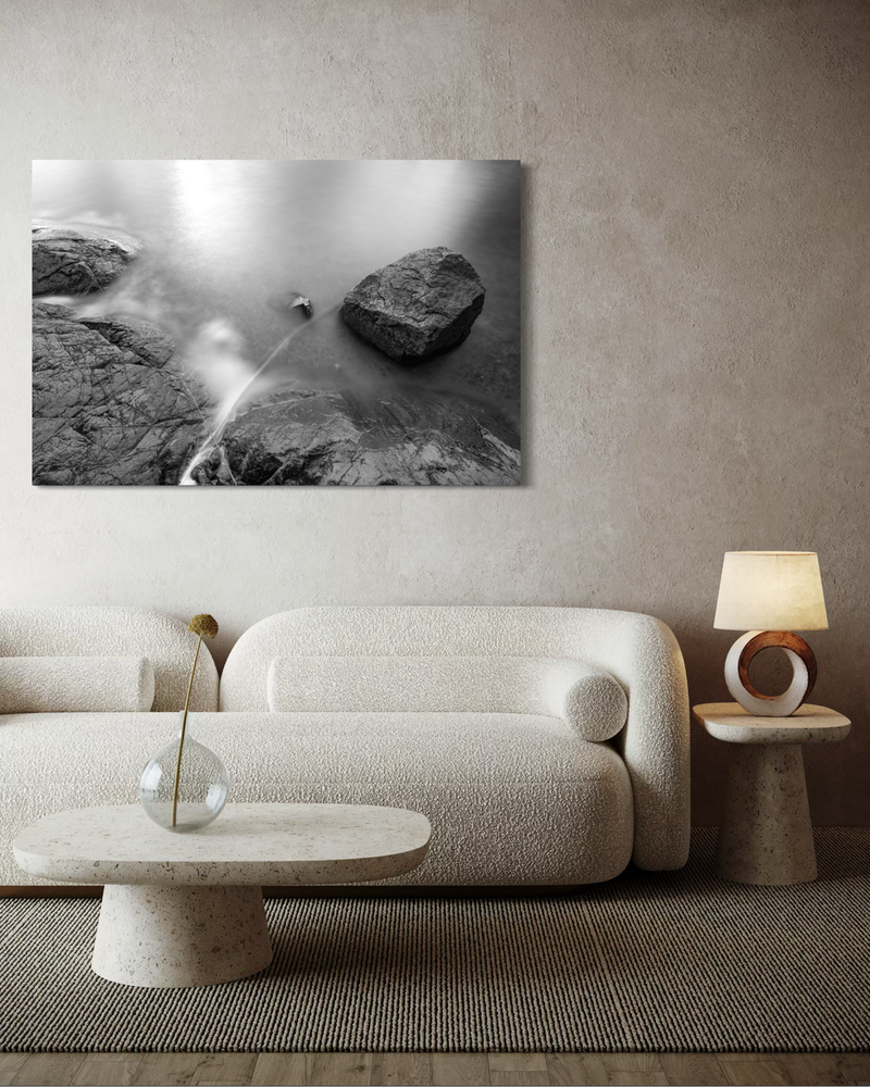 Schwarzweiß-Fotografie auf Leinwand von Klippen und Meer in Langzeitbelichtung, bei der das Wasser sanft um die Felsen fließt und Ruhe ausstrahlt.